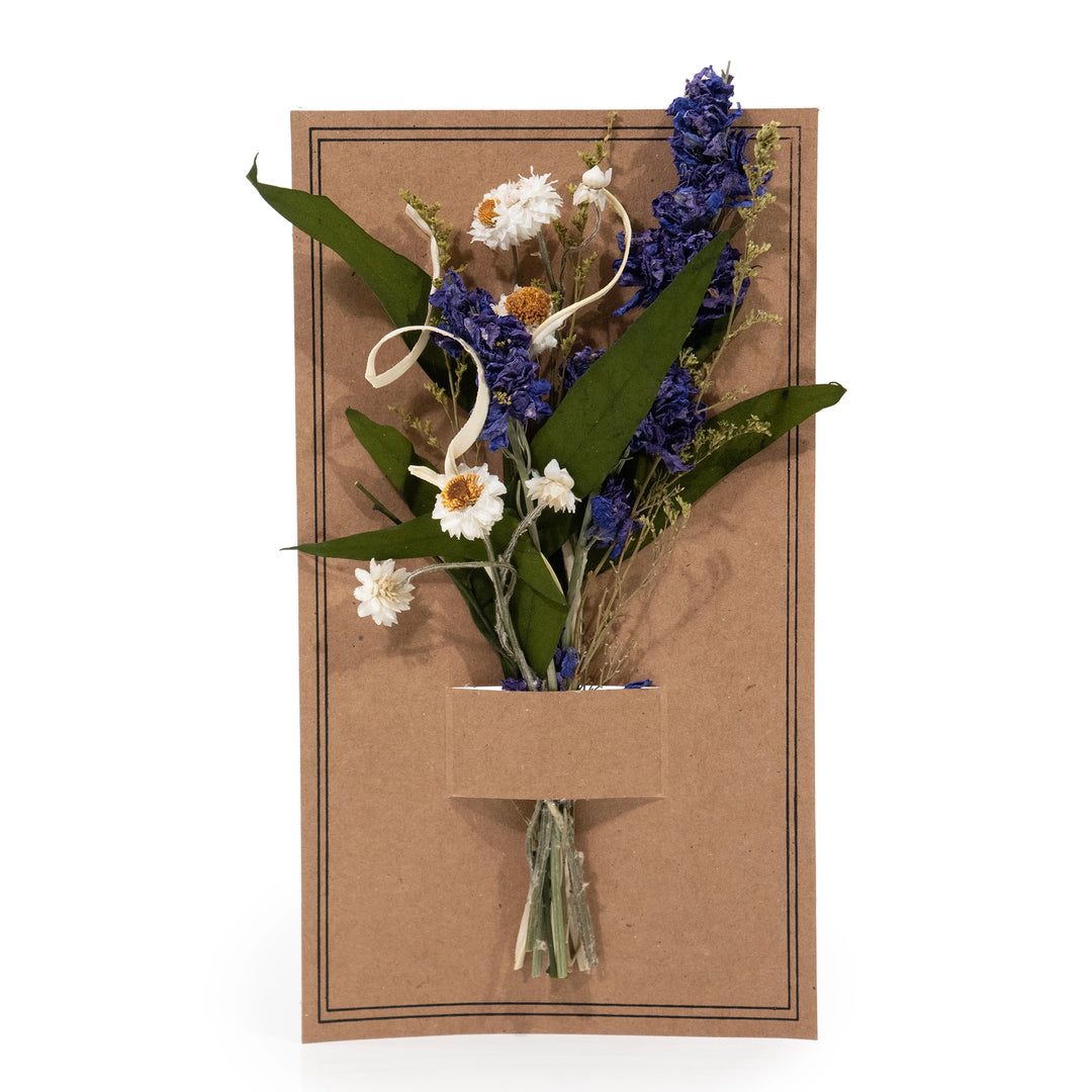 Dried mini bouquet of purple larkspur, white strawflowers, and green eucalyptus leaves with raffia curls, tucked into a pocket on a kraft paper card.