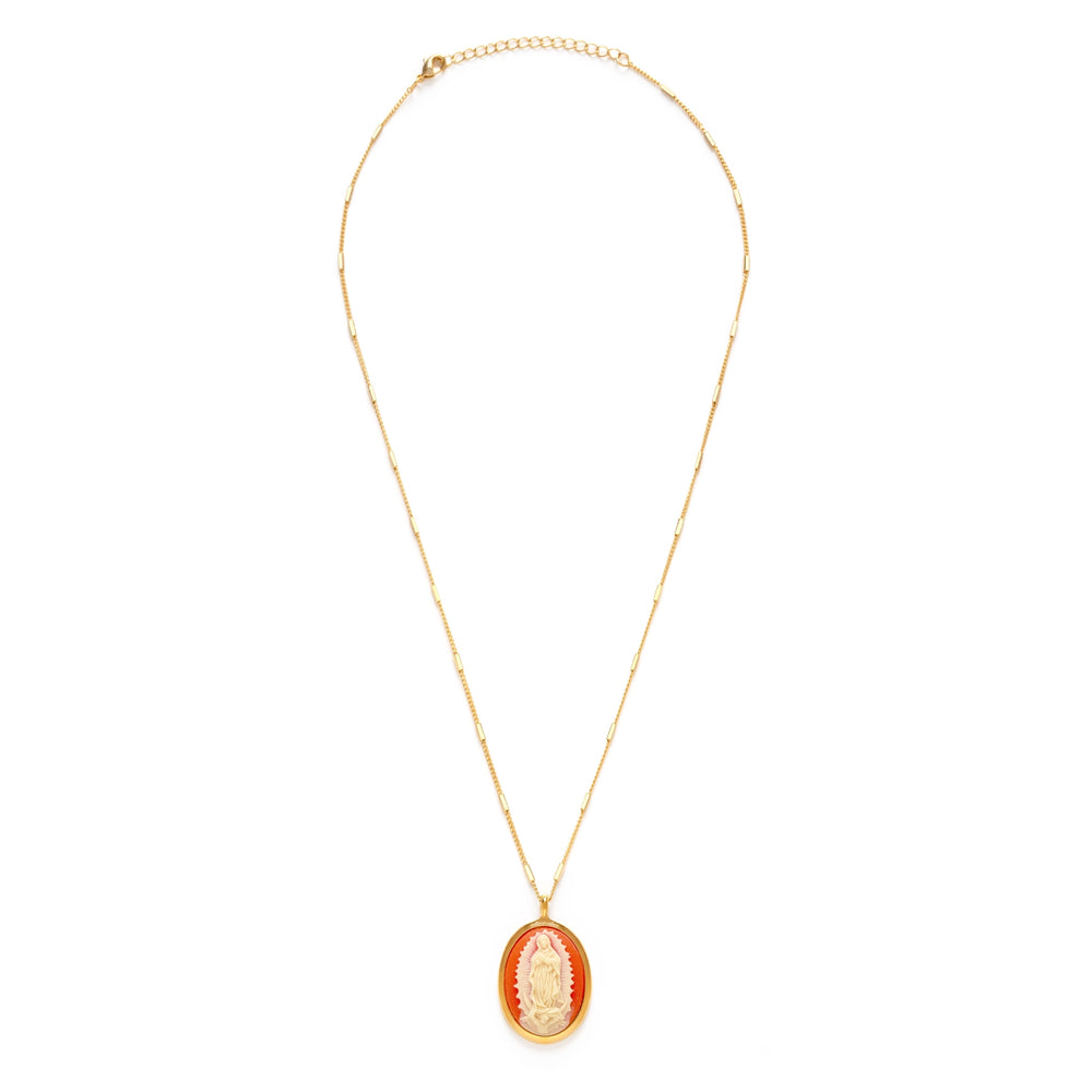 An oval cameo pendant featuring a detailed relief carving of the Virgin of Guadalupe in cream tones against a rich coral background, framed in a polished gold oval bezel, hung on a mixed bar and curb link gold chain.