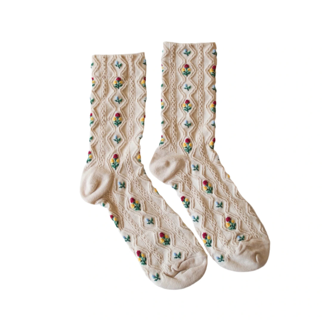 A pair of cream-colored socks with a textured knit pattern and small embroidered flowers in red, yellow, and green arranged in a vertical diamond motif,