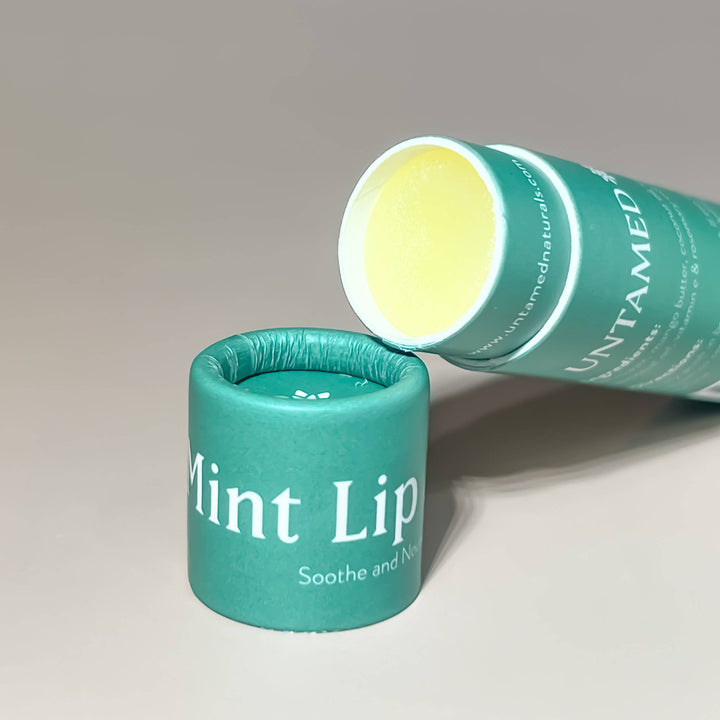 open tube of lip balm laying on the lid. Features green branded label with whtie lettering. Brand: UnTamed Naturals