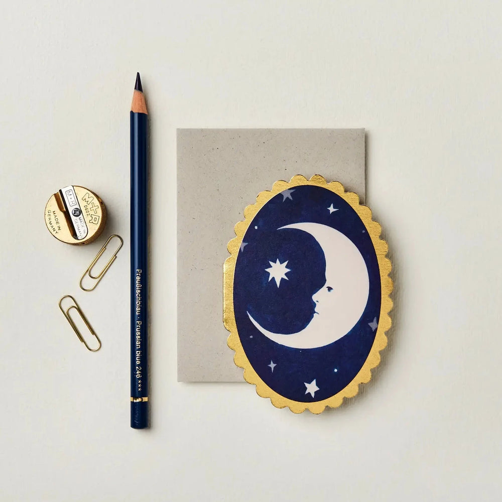 a oval blue card with a gold scalloped edge featuring a crescent moon in the center featuring a blue pencil, two paper clips and a sharpener