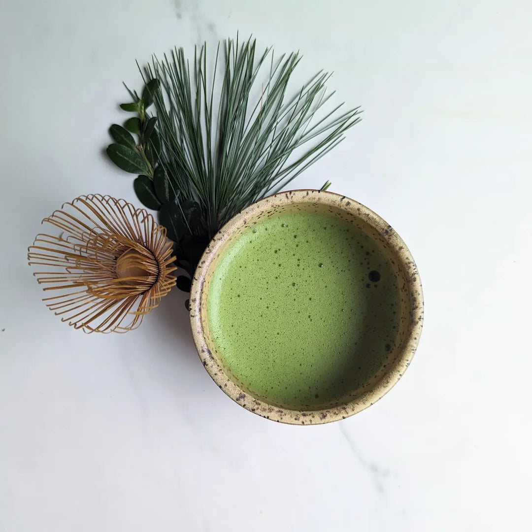 overview of a cup of matcha in a cup with a matcha whisk and green foliage on the side of it.