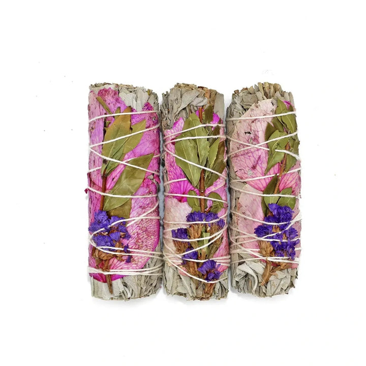 Three sage smudge sticks wrapped with pink rose petals, purple statice flowers, and green leaves, tied with white string