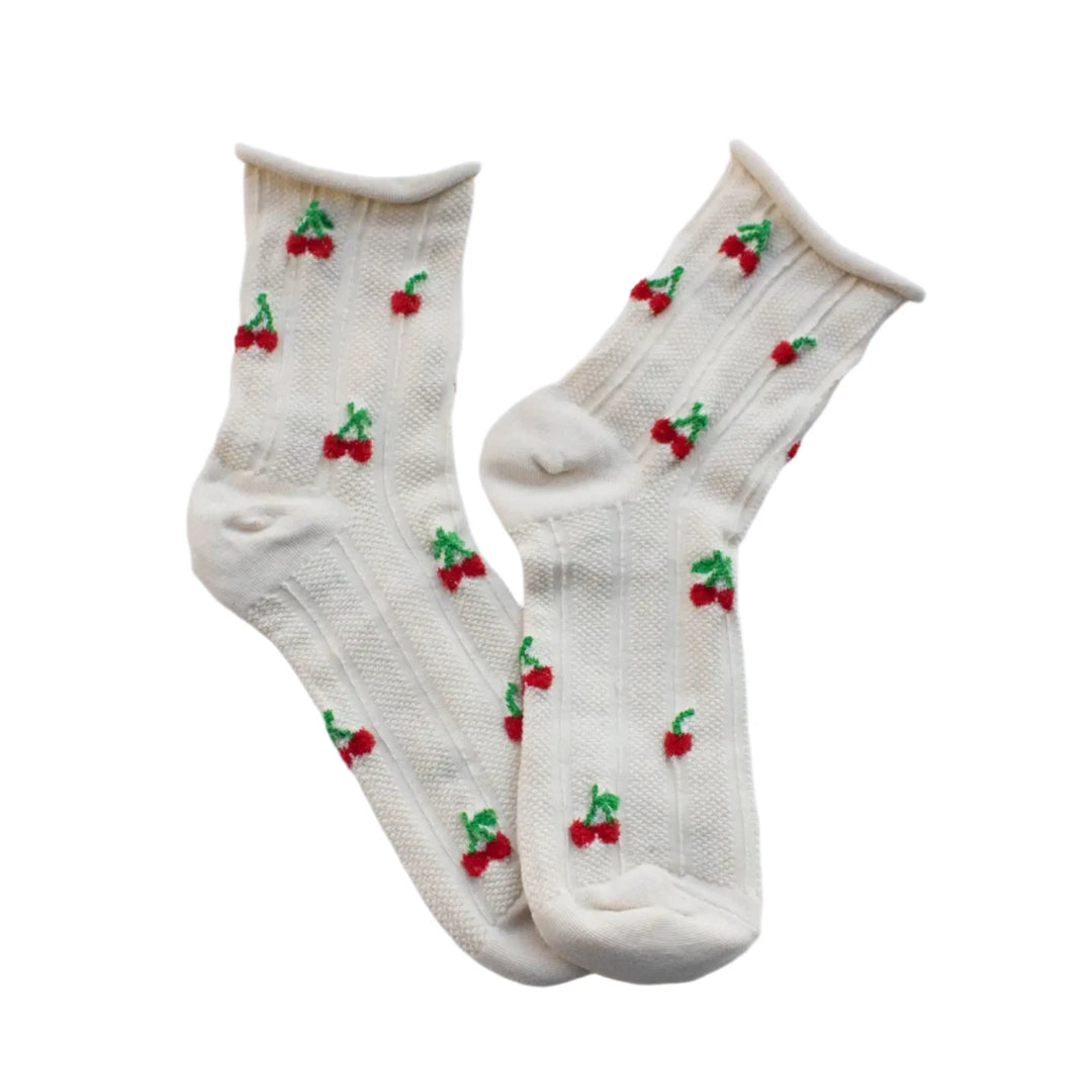 A pair of cream-colored socks with a textured knit design, decorated with small red and green cherry patterns, laid flat.