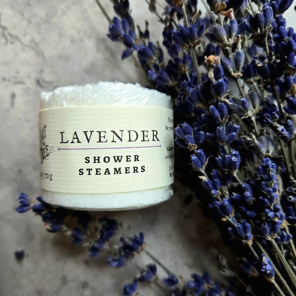 A package of lavender shower steamers wrapped in eco-friendly packaging, placed beside dried lavender sprigs on a textured surface. The label reads "Lavender Shower Steamers," indicating a relaxing and aromatic shower experience.