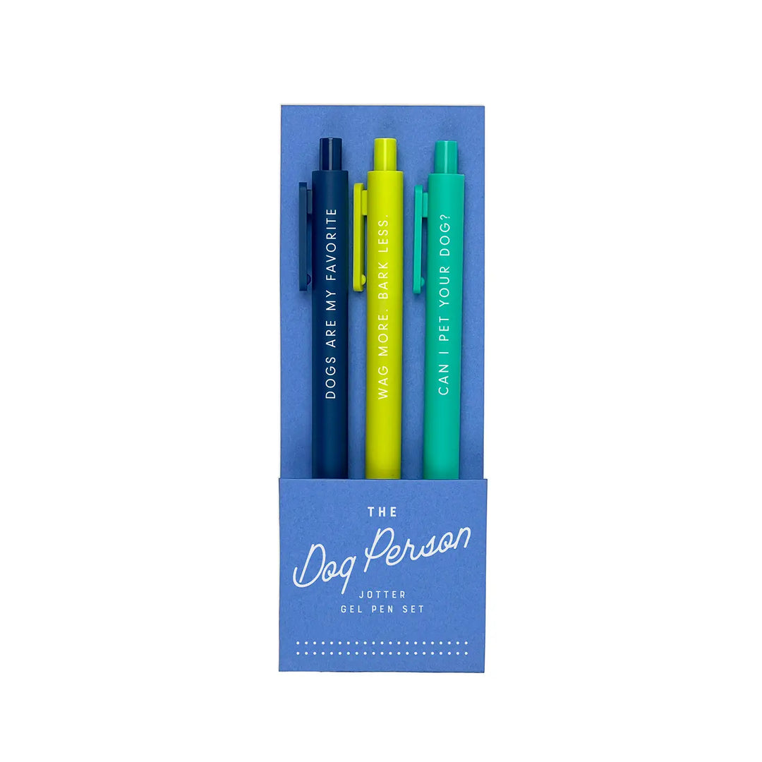 A set of three gel pens in a blue packaging labeled "The Dog Person Jotter Gel Pen Set." The pens come in navy blue, bright yellow, and teal, each featuring a fun dog-themed phrase in white lettering: "Dogs Are My Favorite," "Wag More. Bark Less.," and "Can I Pet Your Dog?"