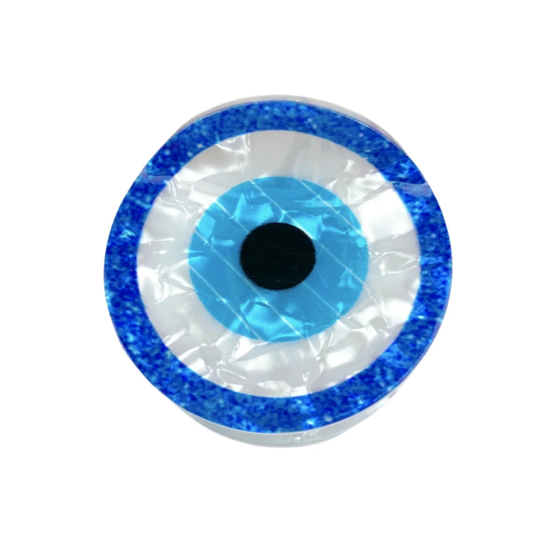 A circular evil eye hair clip with a shimmering blue border, white center, and black pupil, set against a white background.