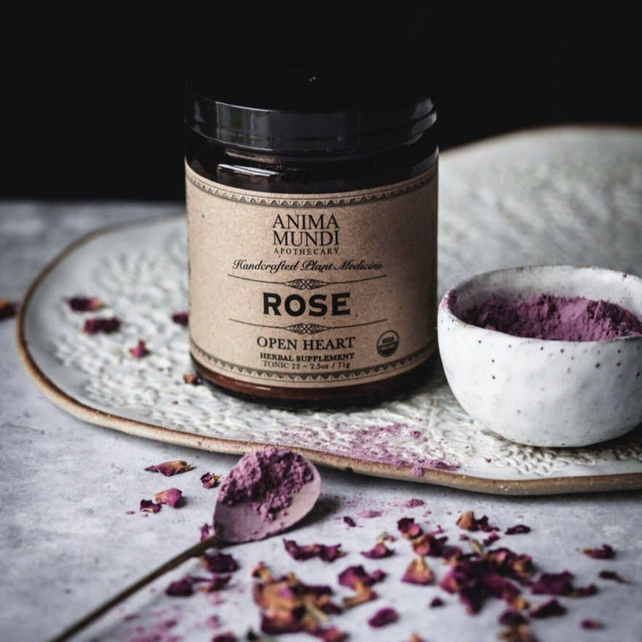 a jar of the rose powder on a dish with a white bowl full of the body to the right of it and a spoon with powder and dried rose petals in front of it. Brand: Anima Mundi Apothecary