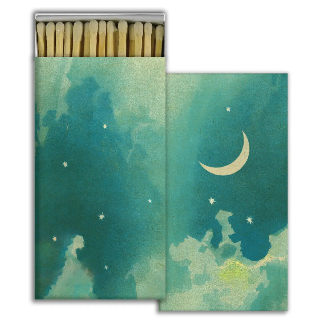 A beautifully designed matchbox featuring a dreamy night sky with shades of blue and green, scattered stars, and a crescent moon. The box is open, revealing long matches with natural wood-tipped ends neatly arranged inside.