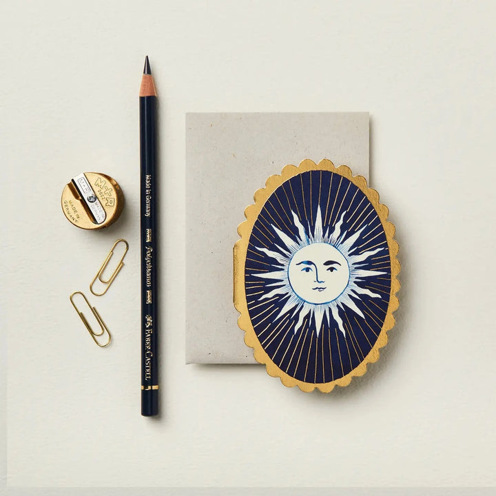 a oval blue card with a gold scalloped edge featuring a sun in the center featurig a black pencil, two paper clips and a sharpener