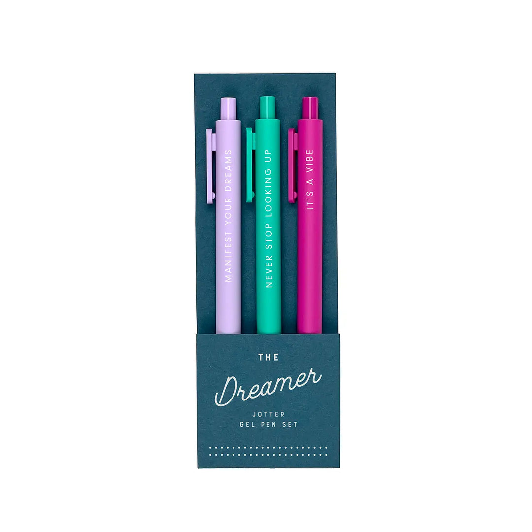 A set of three gel pens in a dark teal packaging labeled "The Dreamer Jotter Gel Pen Set." The pens come in lavender, teal, and magenta, each featuring an uplifting phrase in white lettering: "Manifest Your Dreams," "Never Stop Looking Up," and "It’s a Vibe."