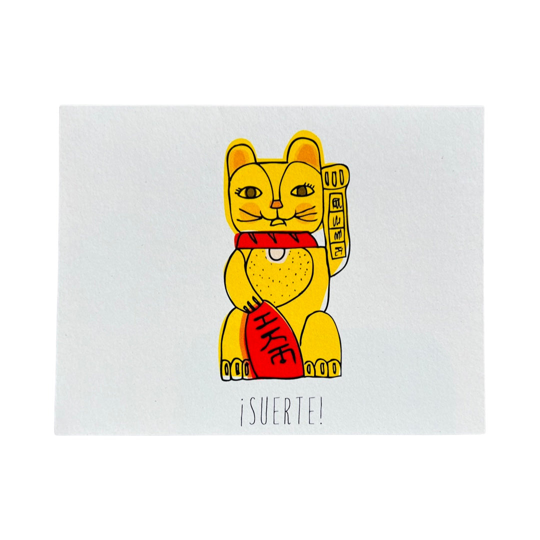 white card with a gold cat with a red object and the word Suerte in black lettering