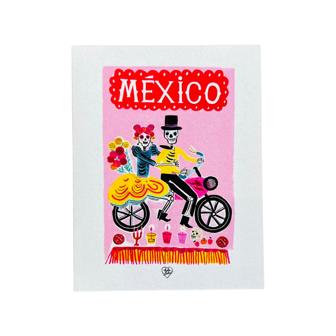 white card with a pink block background and features a calavera couple on a bike with various icons of items found on an altar and the word MEXICO in a papel picado design