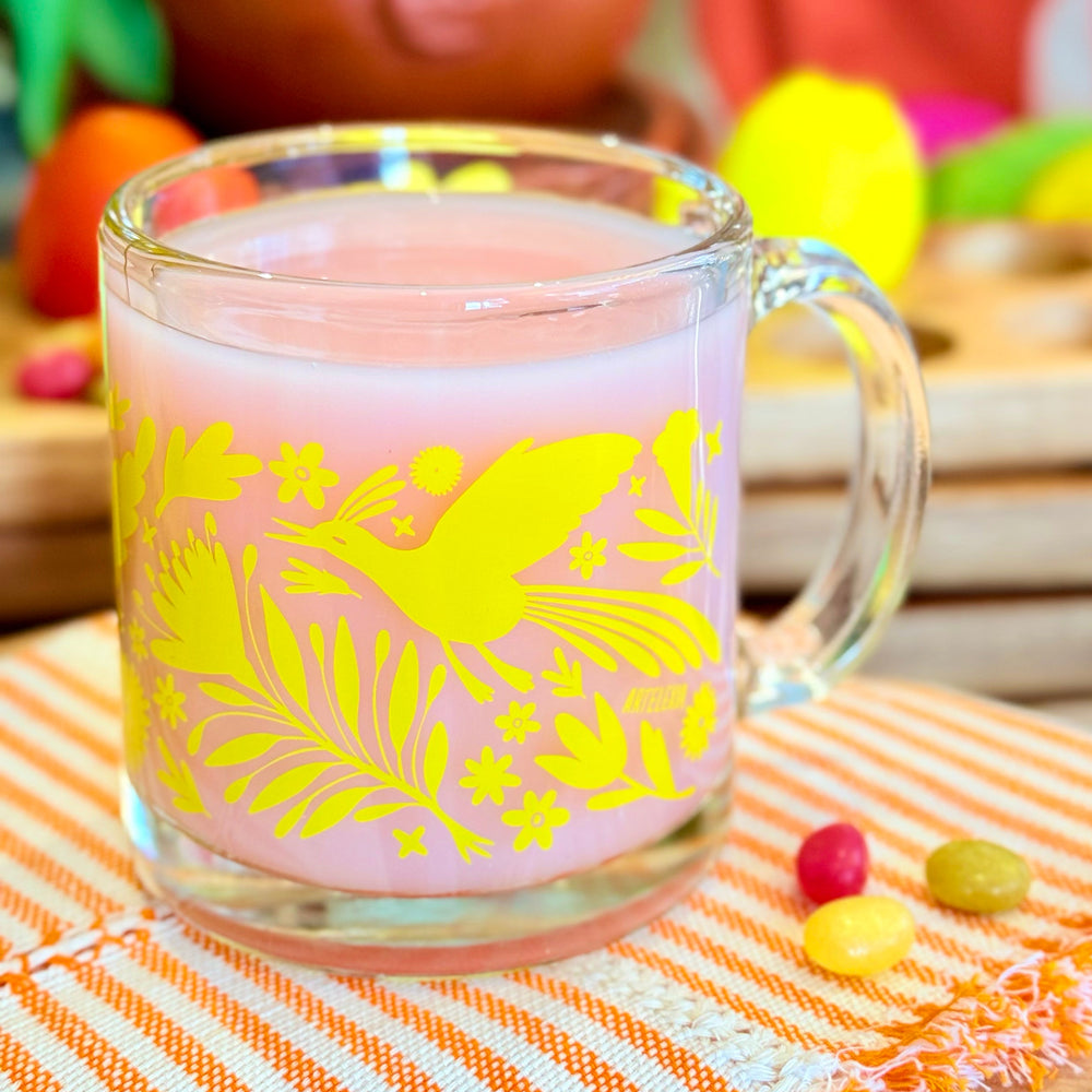 Front view of clear glass mug featuring a wraparound print of a yellow otomi design. The mug is holding a creamy light pink beverage and is sitting on top of an orange and white striped napkin next to a few small candies.