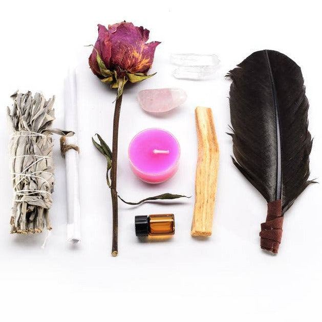 an image of the following items laid out side by side from one another: turkey feather, one pink candle, two crystals, a bottle of jasmine oil, palo santo, dried herbs and rose.