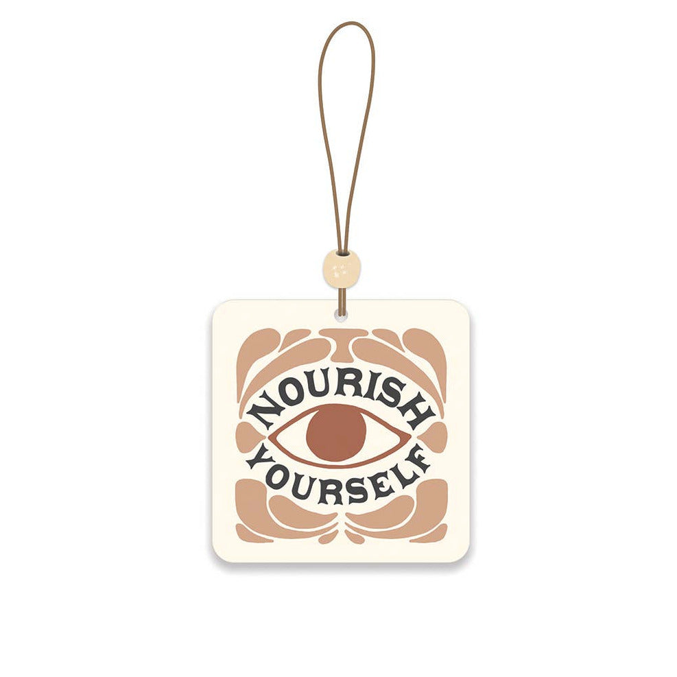 square cream air freshener featuring a brown eye in the center with tan filigree and the phrase Nourish Yourself in black lettering