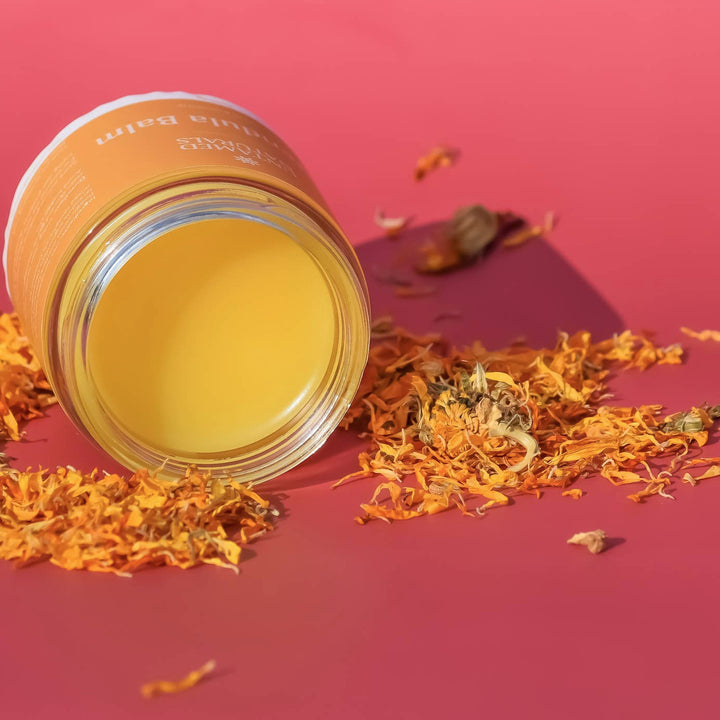 An open jar of calendula balm on its side with a sprinkle of dried calendula petals around it and a pink background. Brand: UnTamed Naturals