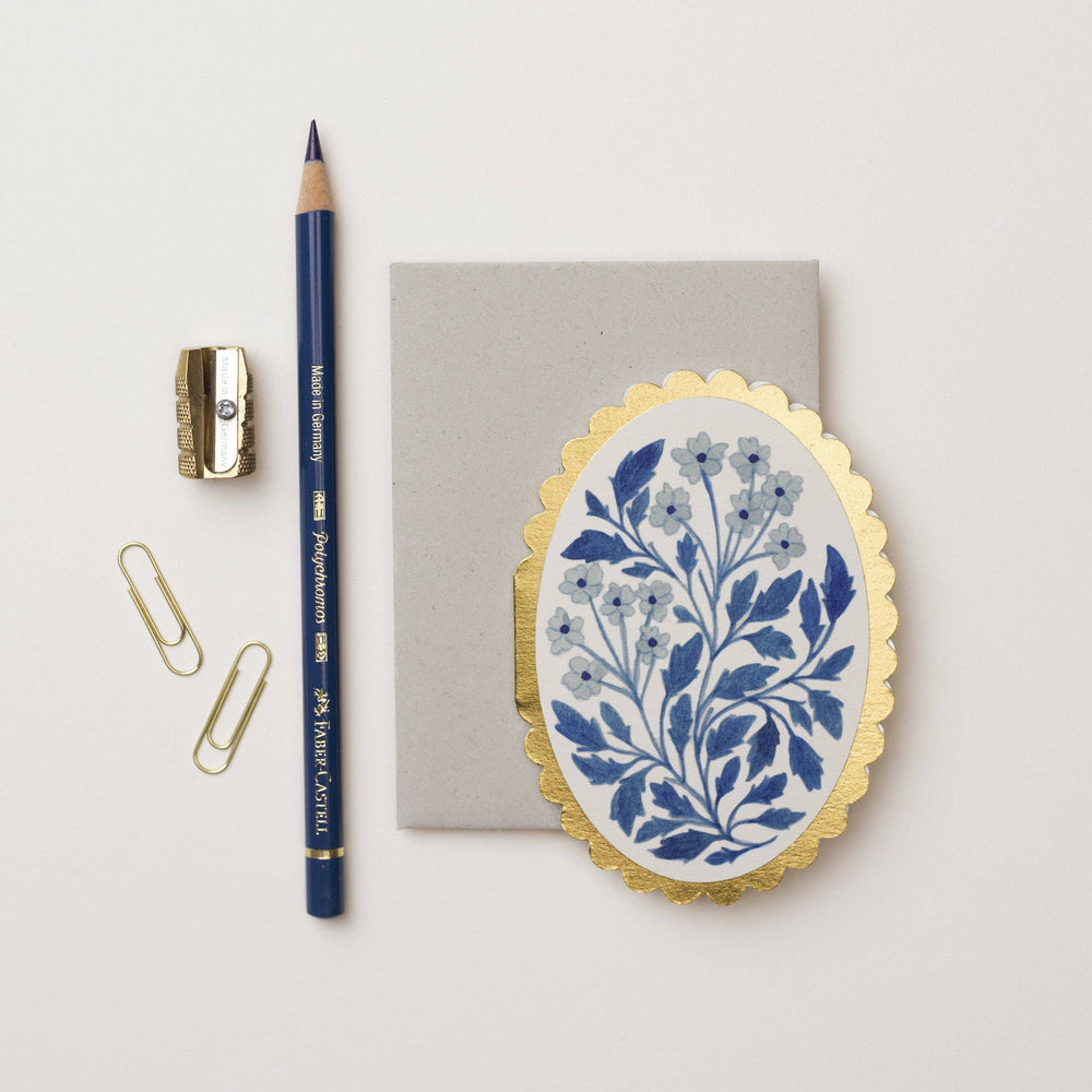oval white card with a gold scalloped edge featuring a blue floral design in the center along side a gray envelope, two paper clips, pencil sharpener and a blue pencil.