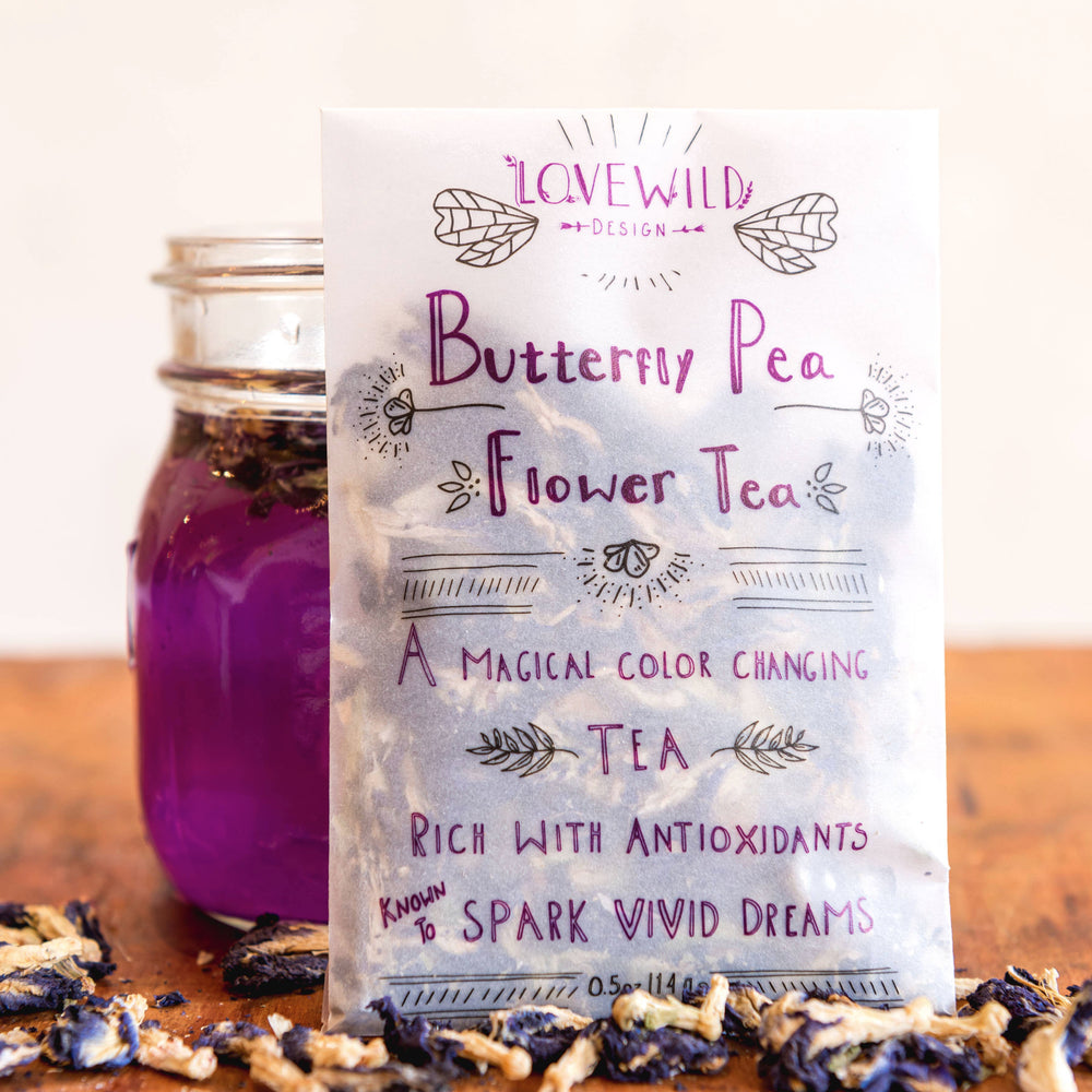 pouch of the butterfly pea flower tea leaning on a glass of the tea and on top of some loose tea. Brand: Lovewild Design